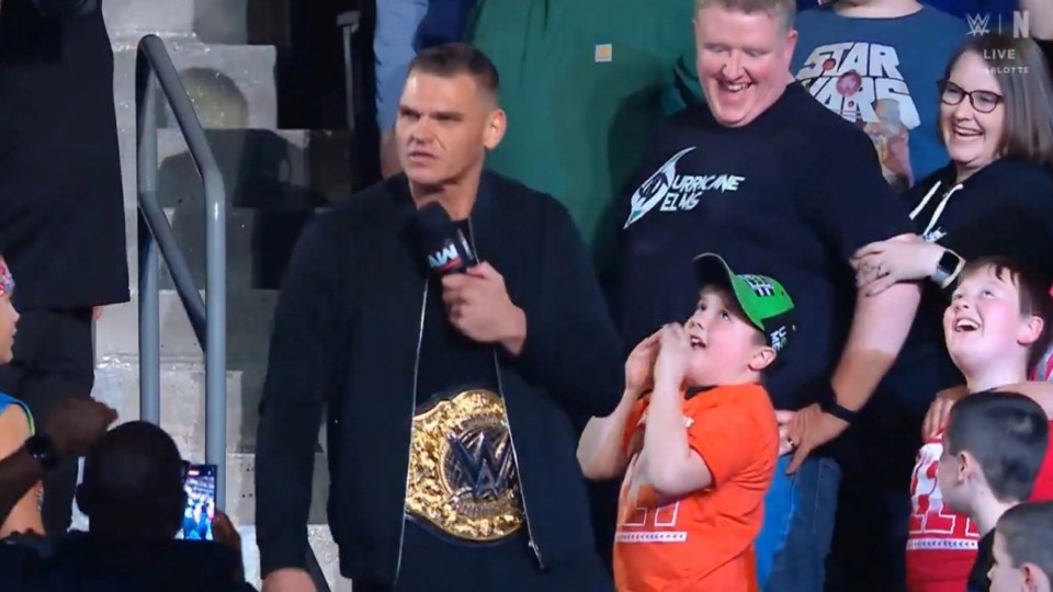 A wrestler wearing a championship belt speaks into a microphone while surrounded by fans.
